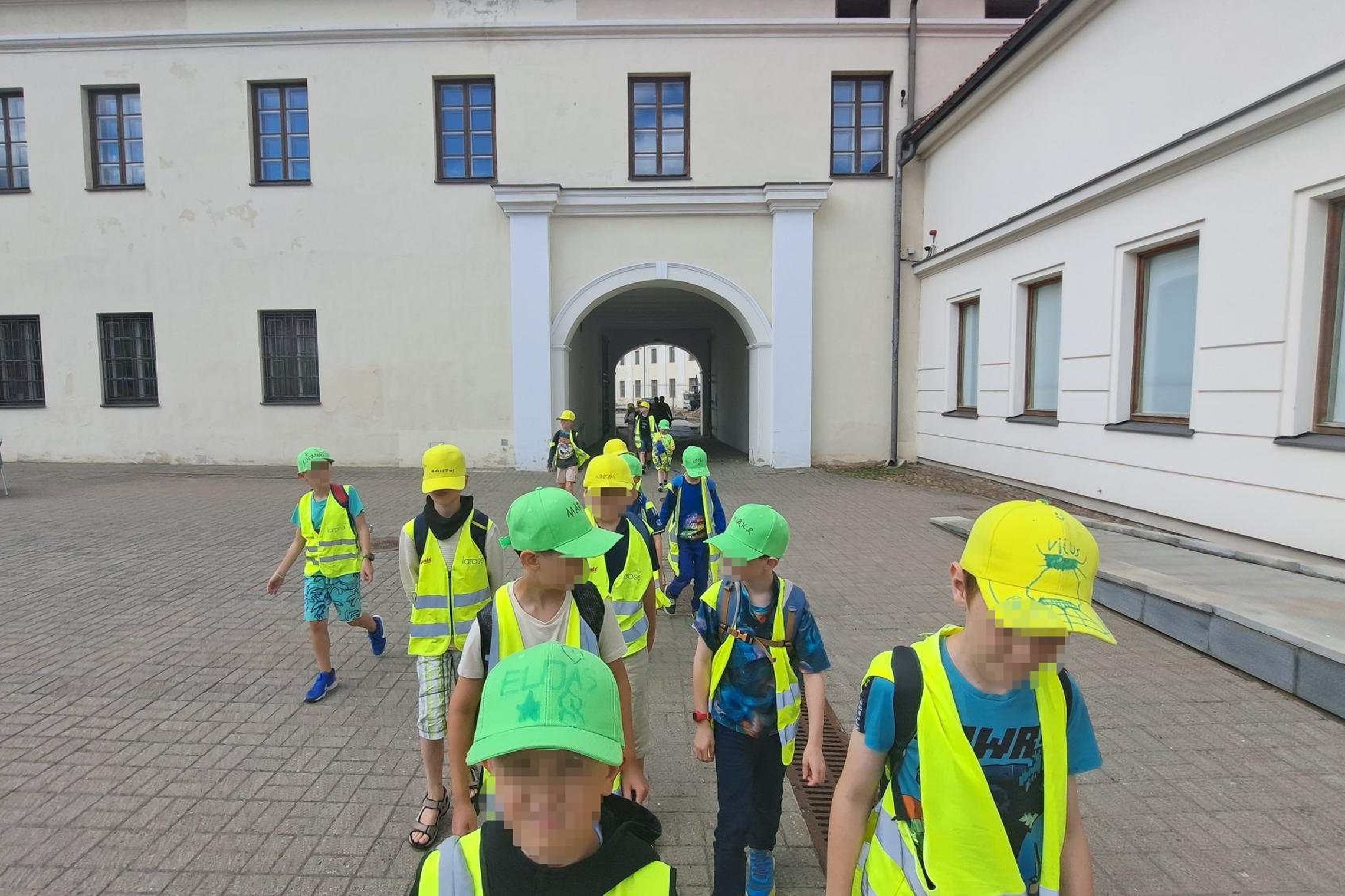 Vaikai su ryškiai geltonomis liemenėmis ir kepuraitėmis žygiuoja žemutinės pilies teritorijoje Vilniuje edukacijos metu.