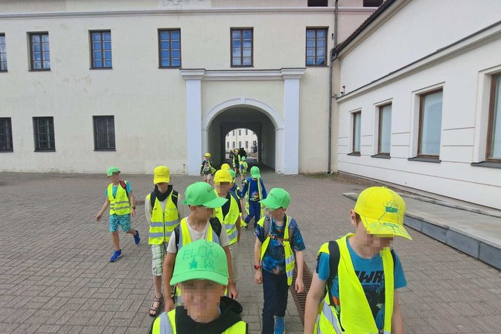 Vaikai su ryškiai geltonomis liemenėmis ir kepuraitėmis žygiuoja žemutinės pilies teritorijoje Vilniuje edukacijos metu.