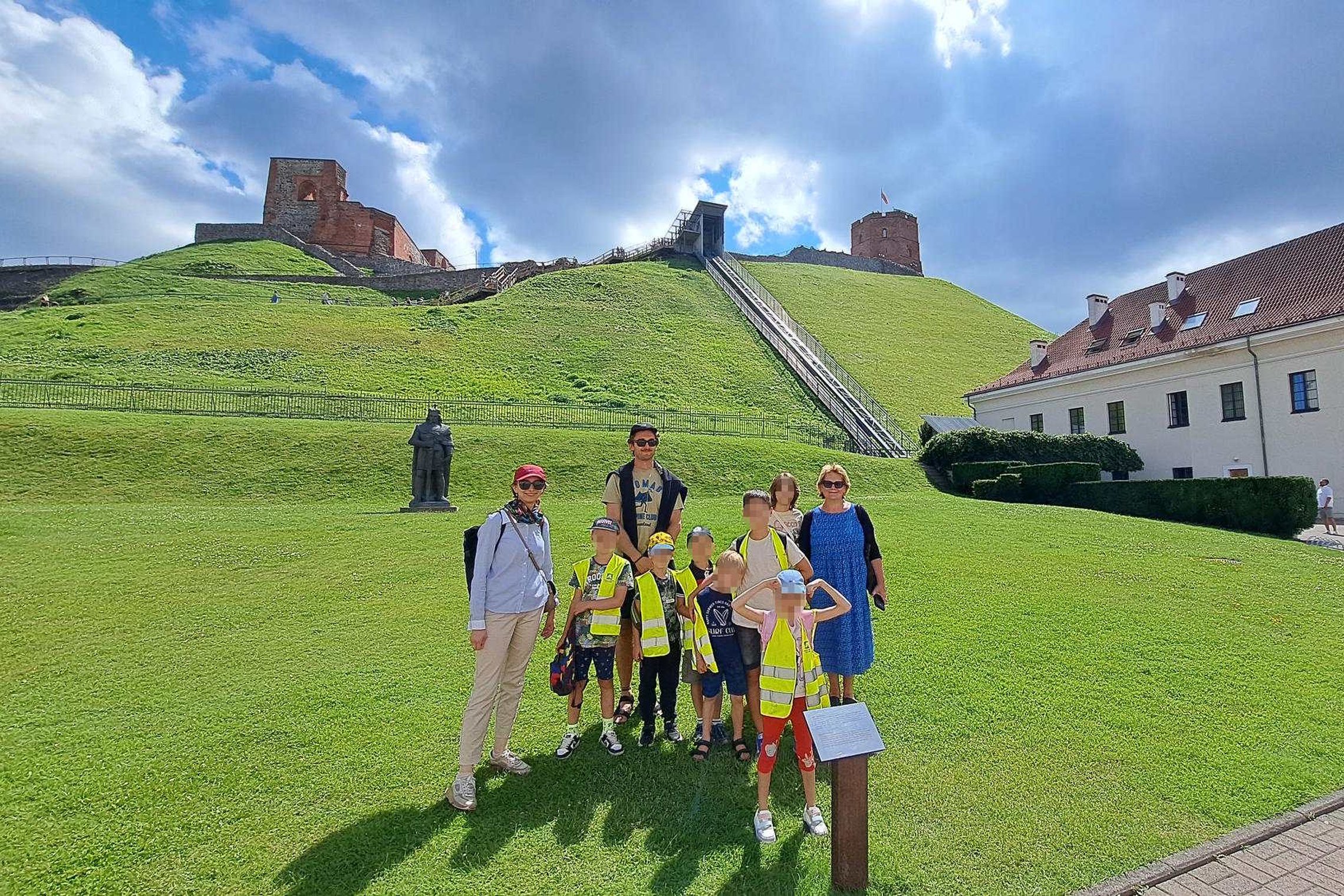 Vaikų grupė su lydinčiaisiais asmenimis pozuoja prie Vilniaus pilių teritorijos, žalioje pievoje priešais Gedimino kalną; fone matomas Gedimino bokštas, Aukštutinės pilies liekanos ir paminklas didžiajam kunigaikščiui Vytautui.