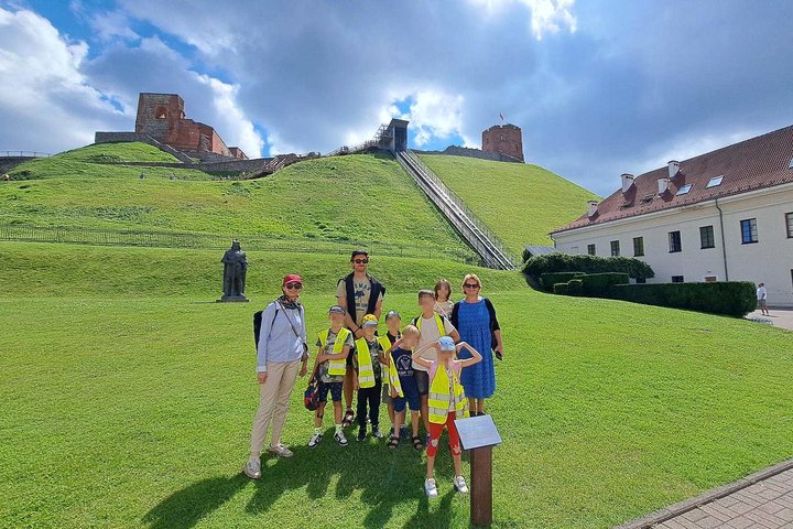 Vaikų grupė su lydinčiaisiais asmenimis pozuoja prie Vilniaus pilių teritorijos, žalioje pievoje priešais Gedimino kalną; fone matomas Gedimino bokštas, Aukštutinės pilies liekanos ir paminklas didžiajam kunigaikščiui Vytautui.