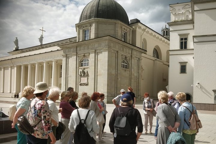 Didelė senjorų ekskursija stovi Vilniaus katedros aišktėje, šalia Gedimino paminklo. Už jų Vilniaus katedra ir Valdovų rūmai.