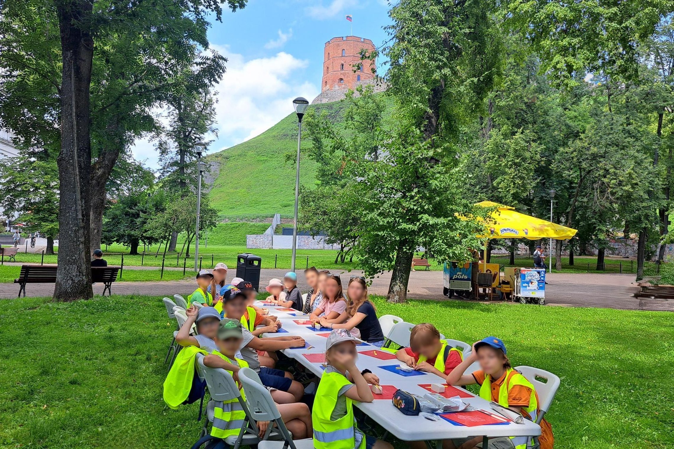 Vaikai sėdi prie ilgų stalų pievoje, Gedimino pilies kalno papėdėje, ir lipdo bokštus edukacijos metu.
