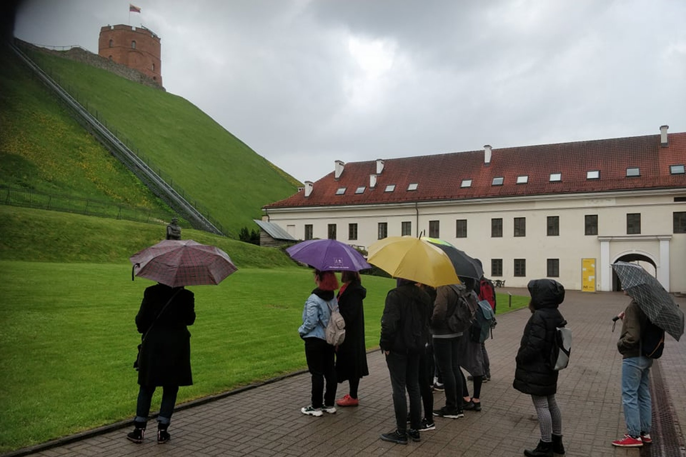 Ekskursijos grupė stovi Geimino kalno papėdėje. Lyja lietus, todėl žmonės yra su lietsargiais. Yra gaivi, nors ir lietinga gegužės diena. Šviežia žole pasidengias kalnas. Nuotraukos gilumoje matosi Lietuvos nacionalinio muziejaus pastatas.