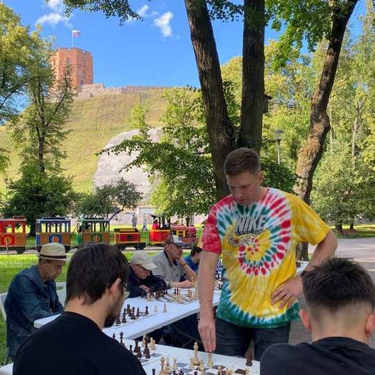 Šachmatų simultanas po atviru dangumi: žaidėjas vaikšto tarp stalų, kur sėdi įvairaus amžiaus dalyviai, o fone stūkso Gedimino pilies bokštas ir važiuoja spalvingas vaikų traukinukas.