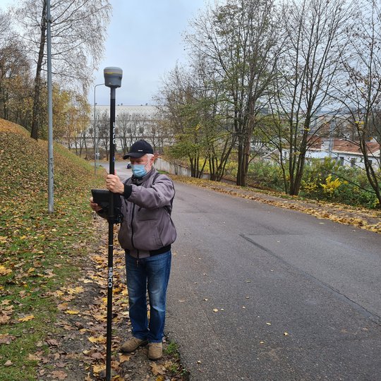 Nuotraukoje žilas žmogus, kuris yra su juodos spalvos kepure, pilkos spalvos striuke, mėlynais džinsais ir smėlio spalvos batais. Jis taip pat užsidėjęs šviesiai mėlynos spalvos veido kaukę. Vyras rankose laiko juodos spalvos matavimo prietaisą. Už jo deš