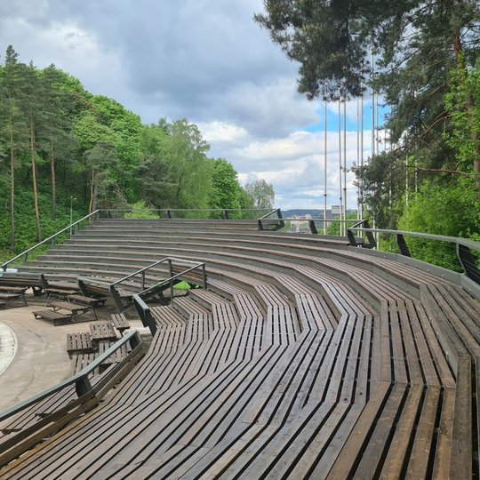 Dainų slėnio suremontuota estrada. Gražūs, nauji mediniai suolai. Yra vasara, žaliuoja medžiai.