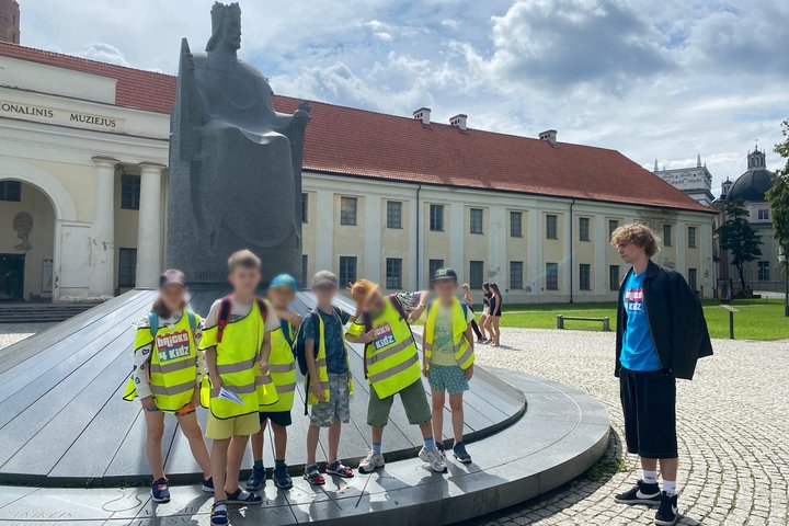 Vaikai su vadovu pozuoja prie Mindaugo paminklo šalia Nacionalinio muziejaus Vilniuje – visi dėvi ryškias saugos liemenes, aplink matyti istorinis pastatas ir turistai