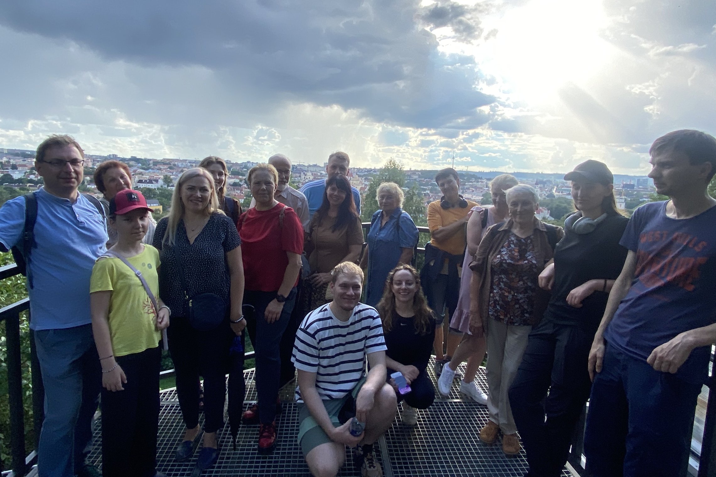 Grupė ekskursijos dalyvių po pasakojimo apie Janiną Dłuską stovi ant Altanos bokštelio, už jų atsiveria įspūdinga Vilniaus panorama su debesuotu dangumi ir pro debesis besiskverbiančia saulės šviesa