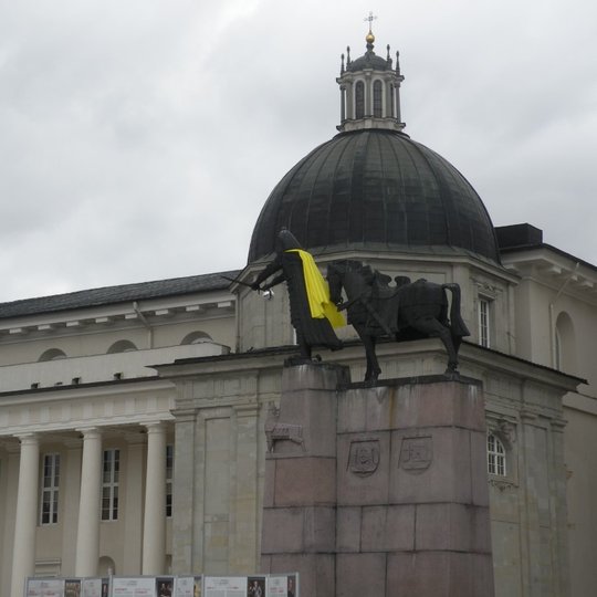 Nuotraukoje iš tolo matoma Katedros aikštė. Nuotraukos centre yra Vilniaus katedra ir metalinė Gedimino ir žirgo statula - paminklas ant aukšto betoninio pagrindo. Gediminas yra su šviesiu apsiaustu. Lauke apsiniaukę ir debesuota, taip pat ir vėjuota, nes