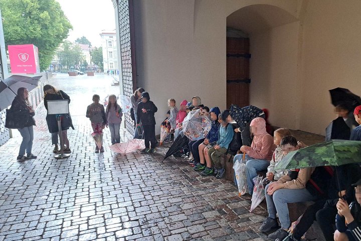 Vaikai su skėčiais ir lietpalčiais glaudžiasi po arkada prie Valdovų rūmų įėjimo, saugodamiesi nuo lietaus edukacijos metu.