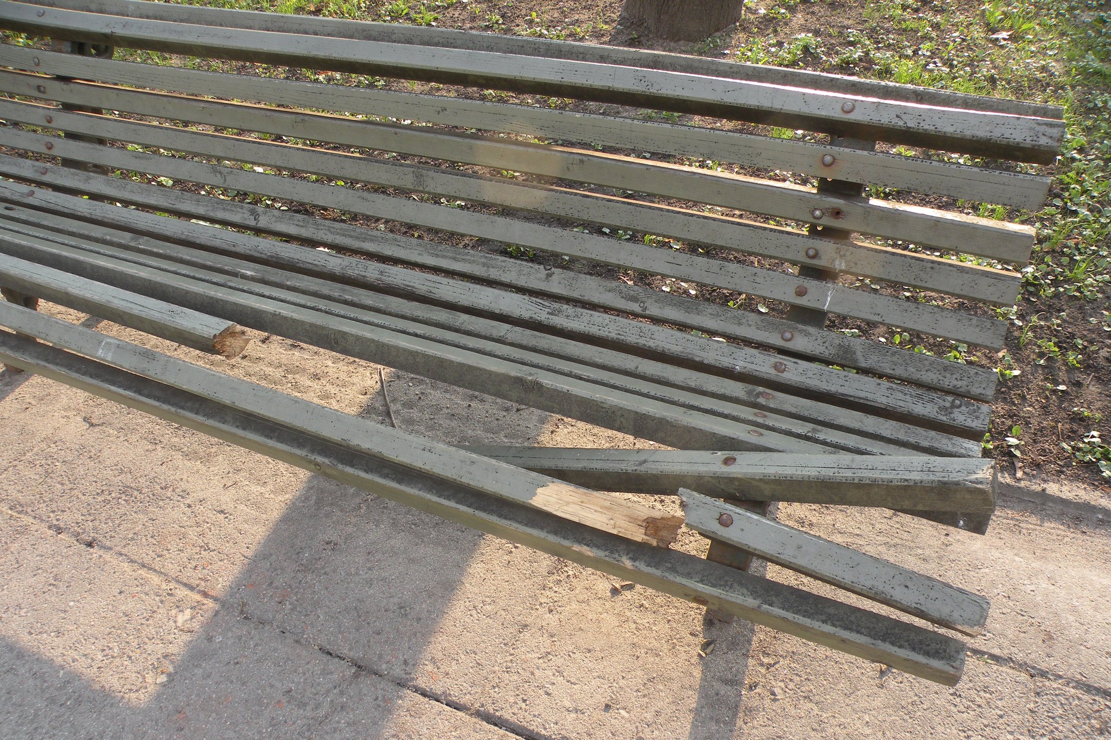 Sulaužytas suoliukas Pilių parke. Jis yra medinis ir iš kelių lentelių. Kelios lentos yra sulaužytos.