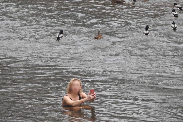 Nuotraukoje upė Vilnelė ir panirus vandenyje iki pečių jauna, šviesių plaukų moteris. Ji bando nusifotografuoti save su mobiliu telefonu. Laiko savo rankose telefoną, bet yra pasisukusi į dešinę, žiūri į krantą, nes mato, kad ją fotografuoja. Ji nepyksta