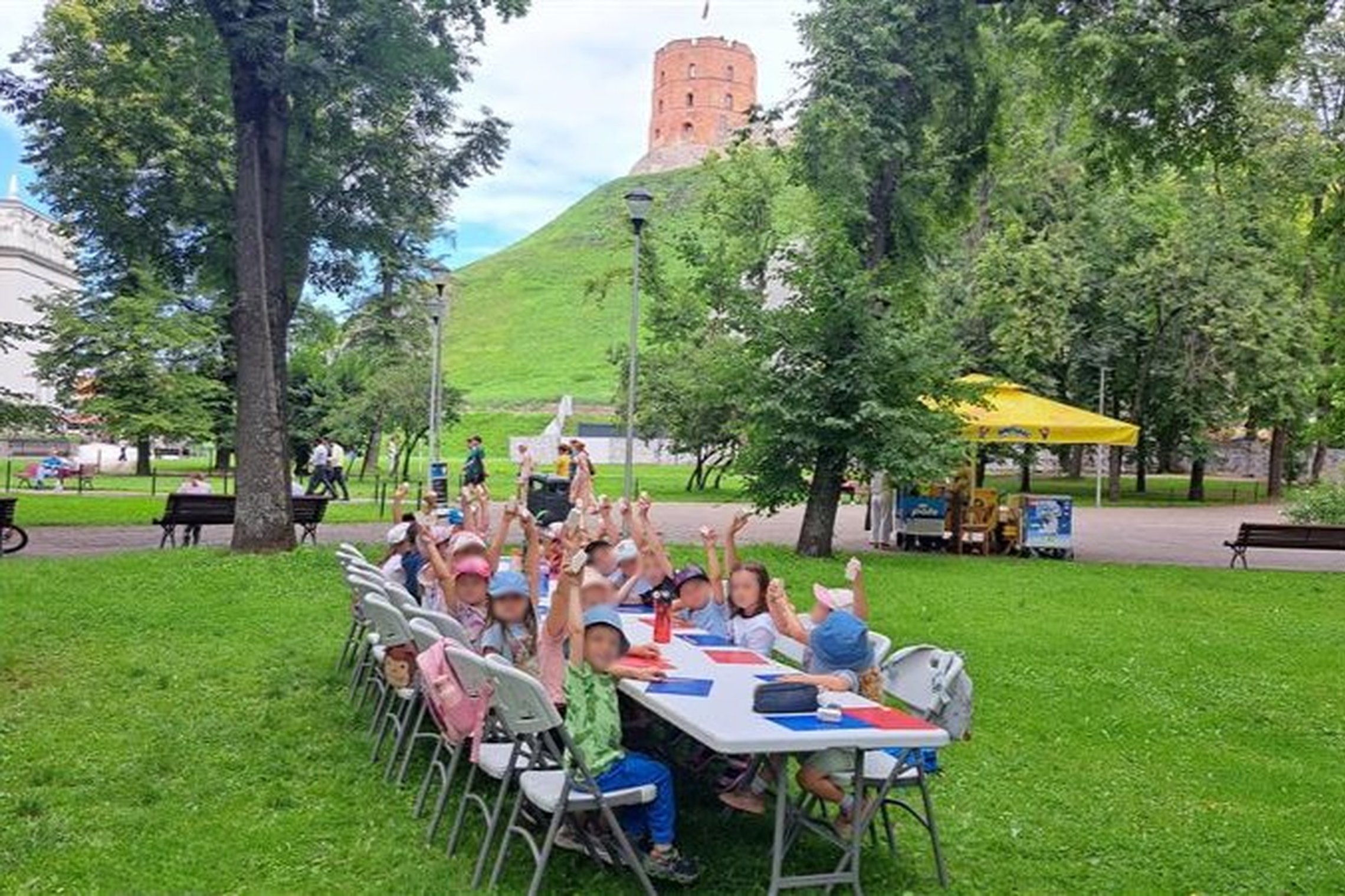 Vaikai sėdi lauke, prie stalo, pakėlę rankas rodo savo nulipdytus bokštus, tolumoje matyti Gedimino pilies bokštas.