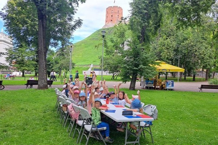 Vaikai sėdi lauke, prie stalo, pakėlę rankas rodo savo nulipdytus bokštus, tolumoje matyti Gedimino pilies bokštas.