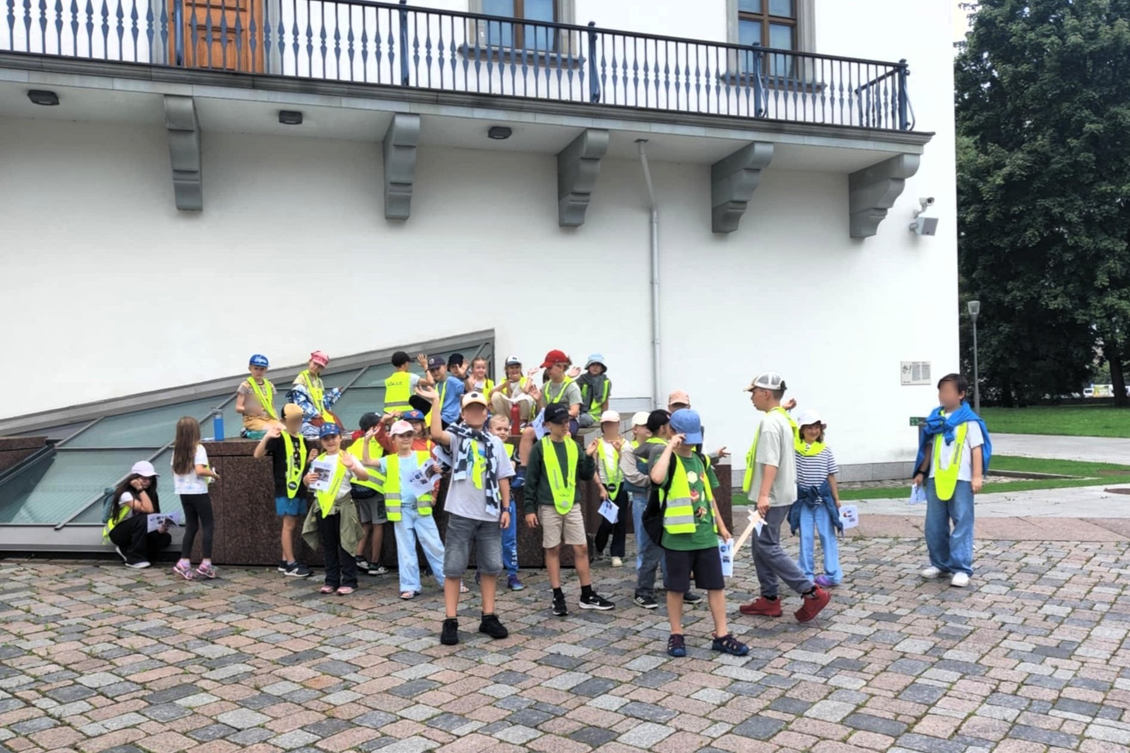 ŠVaikų grupė su suaugusiuoju, dalyvauja edukacinėje veikloje. Jie dėvi ryškiaspalves liemenes su atšvaitais – tai rodo, kad tai organizuota grupė, stovyklautojai. Vaikai stovi prie stiklinio stogo šalia balto pastato su balkonėliu ir saugos kameromis. Gri