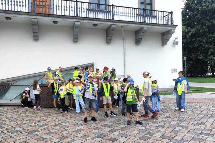 ŠVaikų grupė su suaugusiuoju, dalyvauja edukacinėje veikloje. Jie dėvi ryškiaspalves liemenes su atšvaitais – tai rodo, kad tai organizuota grupė, stovyklautojai. Vaikai stovi prie stiklinio stogo šalia balto pastato su balkonėliu ir saugos kameromis. Gri