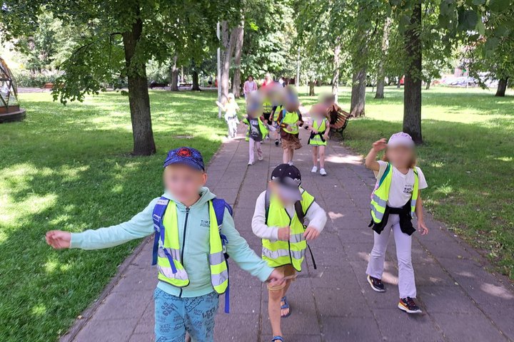 Vaikų grupė su ryškiaspalvėmis liemenėmis eina parko takeliu ir mostais vaizduoja, kad plaukia.