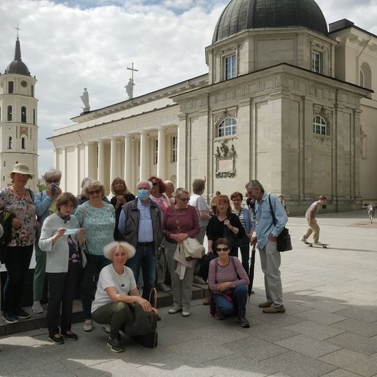 Didelė senjorų ekskursija stovi Vilniaus katedros aišktėje, šalia Gedimino paminklo. Už jų Vilniaus katedra.