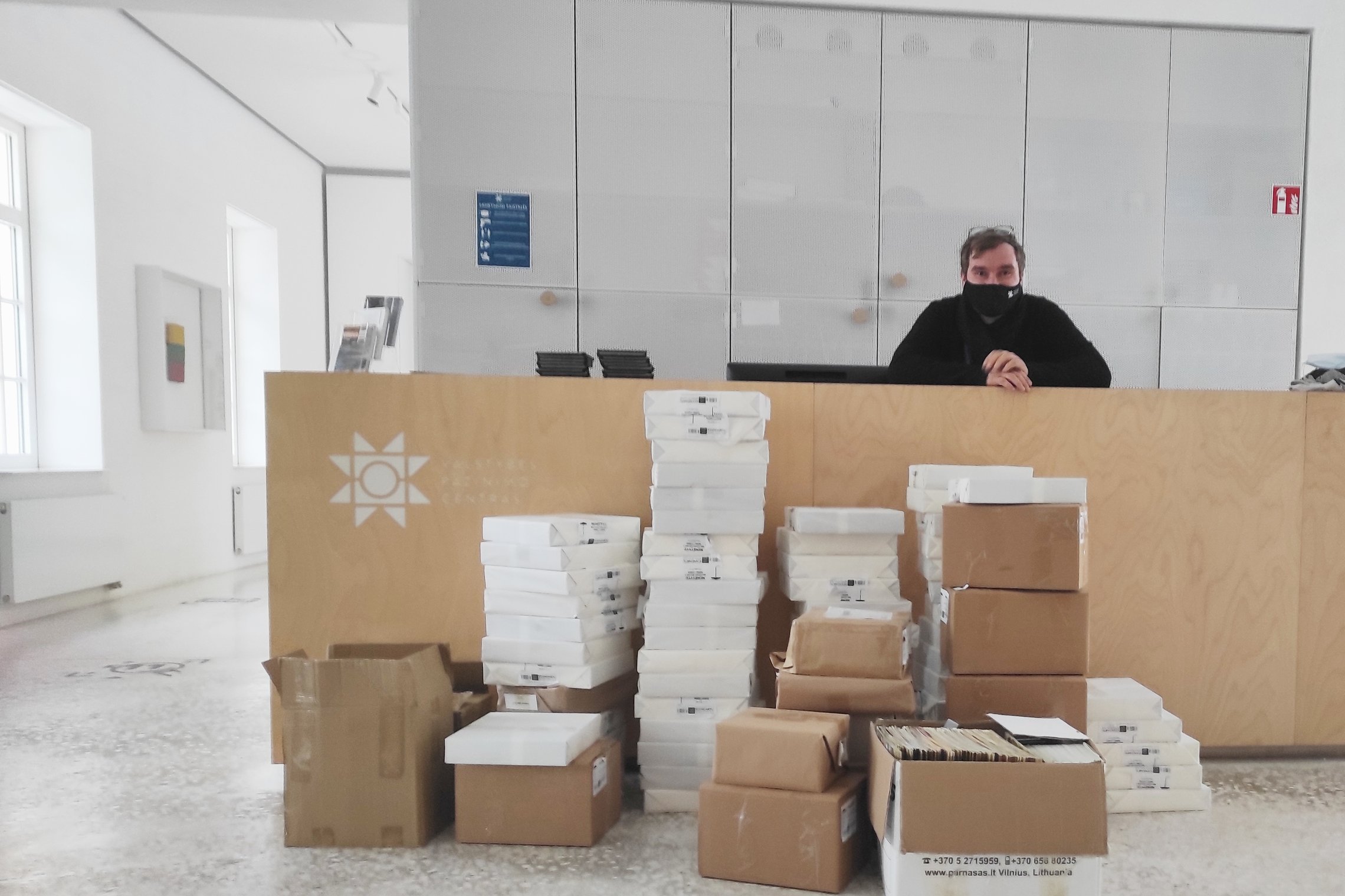  Priekyje centre sukrautos kartoninės dėžės. Visi paketai sudėti nelabai tvarkingai, atsikišusiais kampais, pasikreipę.  Už minėtų dėžių arčiau informacinio centro budėtojo atitvaro regime ant kažko padėtus du paketus, o už baltos dėžės aiškiai regime tri