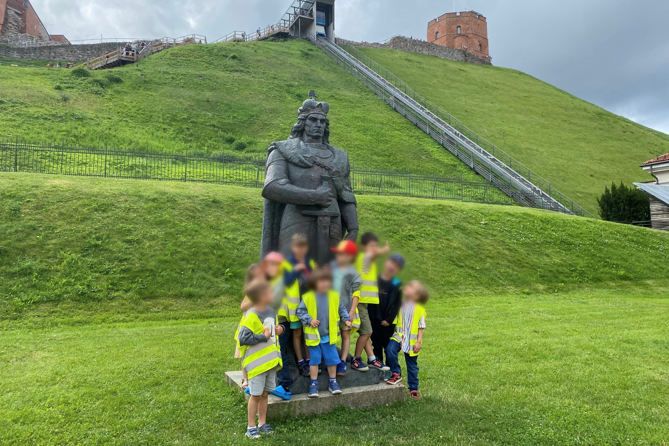 „Bricks4Kids“ stovyklos vaikai prie Gedimino skulptūros, už jų – žalias kalnas ir pilies bokštas