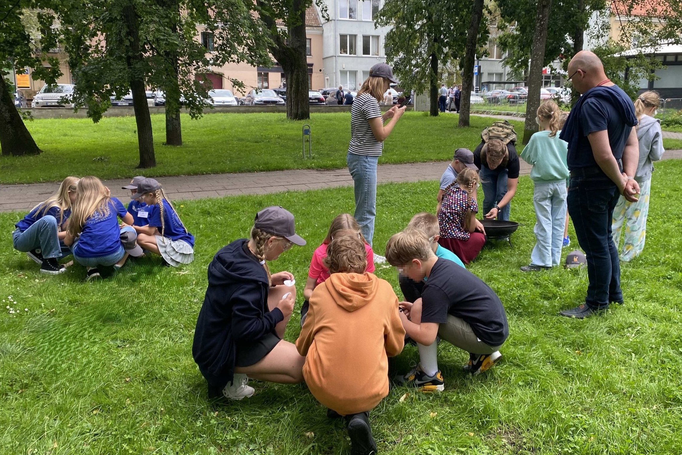 Vaikai susiskirstę į grupeles pievoje dalyvauja edukacijoje apie ugnį. Vieni stebi ugnies kūrimo priemones, kiti diskutuoja tarpusavyje. Aplink vaikšto vadovai ir stebi.