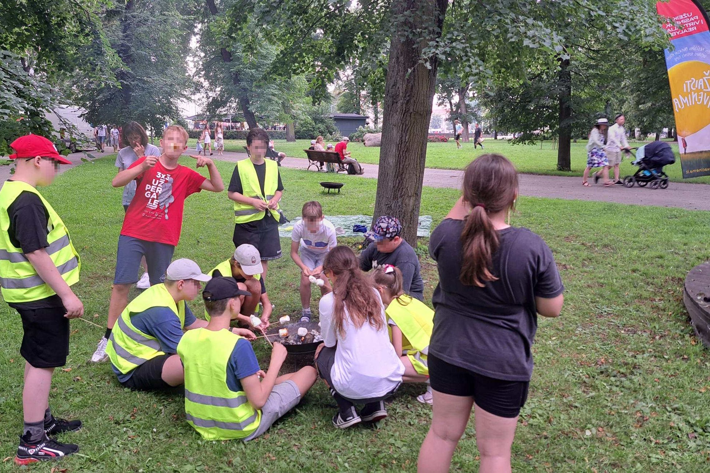 Grupė vaikų su ryškiaspalvėmis saugos liemenėmis ratu aplink nedidelę laužavietę parke kepa zefyrus – Tauro sporto mokyklos vasaros stovyklos Ugnies edukacijos akimirka