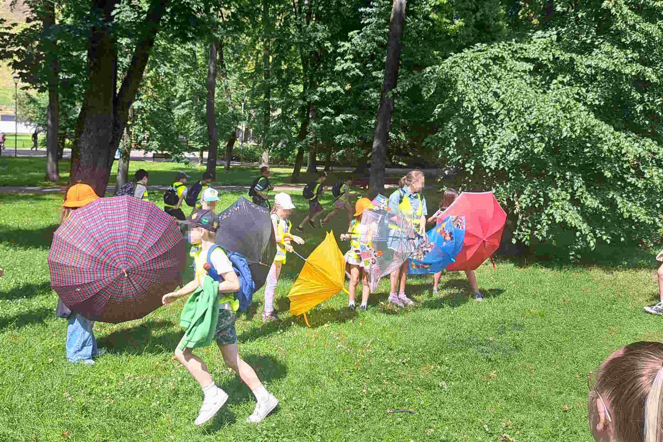 Vaikai su spalvotais skėčiais ir kuprinėmis žaidžia žalioje pievoje tarp medžių Vilniaus pilių teritorijoje, vasaros stovyklos metu.
