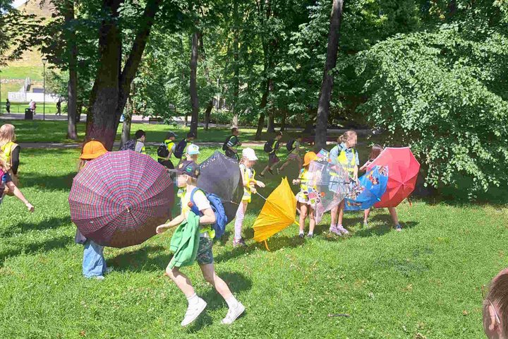Vaikai su spalvotais skėčiais ir kuprinėmis žaidžia žalioje pievoje tarp medžių Vilniaus pilių teritorijoje, vasaros stovyklos metu.
