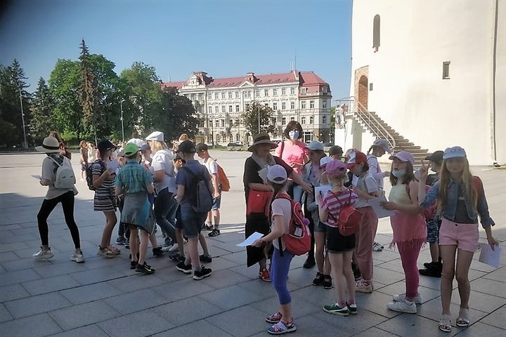 Didelė vaikų grupė stovi šalia Vilniaus katedros varpinės. Vaikai su vasarinėmis kepurėmis, su kuprinėmis ir užduočių lapais. Vyksta orientacinis žaidimas.