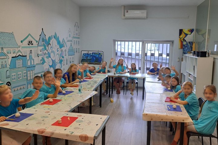 Nuotraukoje vaikai sėdi prie stalų gražioje klasėje. Klasės sienos puoštos piešiniais.