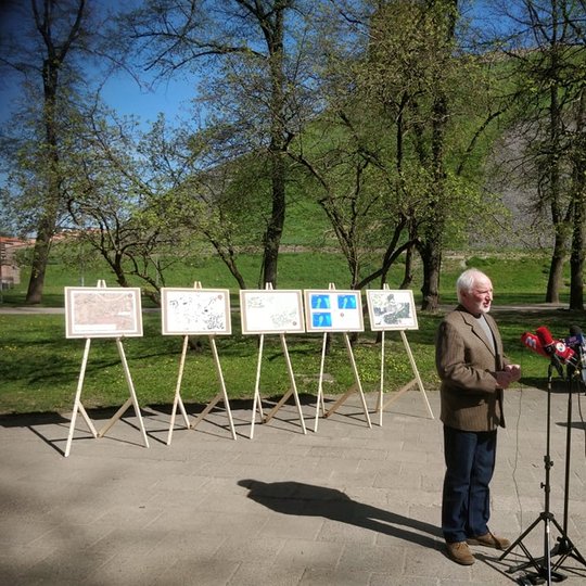 Pilių parkas, vyksta spaudos konferencija. Išrikiuoti penki molbertai su spalvotais plakatais, kuriuose vaizduojami atlikti tyrimai randant bokštą. Už molbertų medžiai, o už jų Gedimino kalnas ir Gedimino pilies bokštas. Prieš molbertus stovi žmogus. Prie