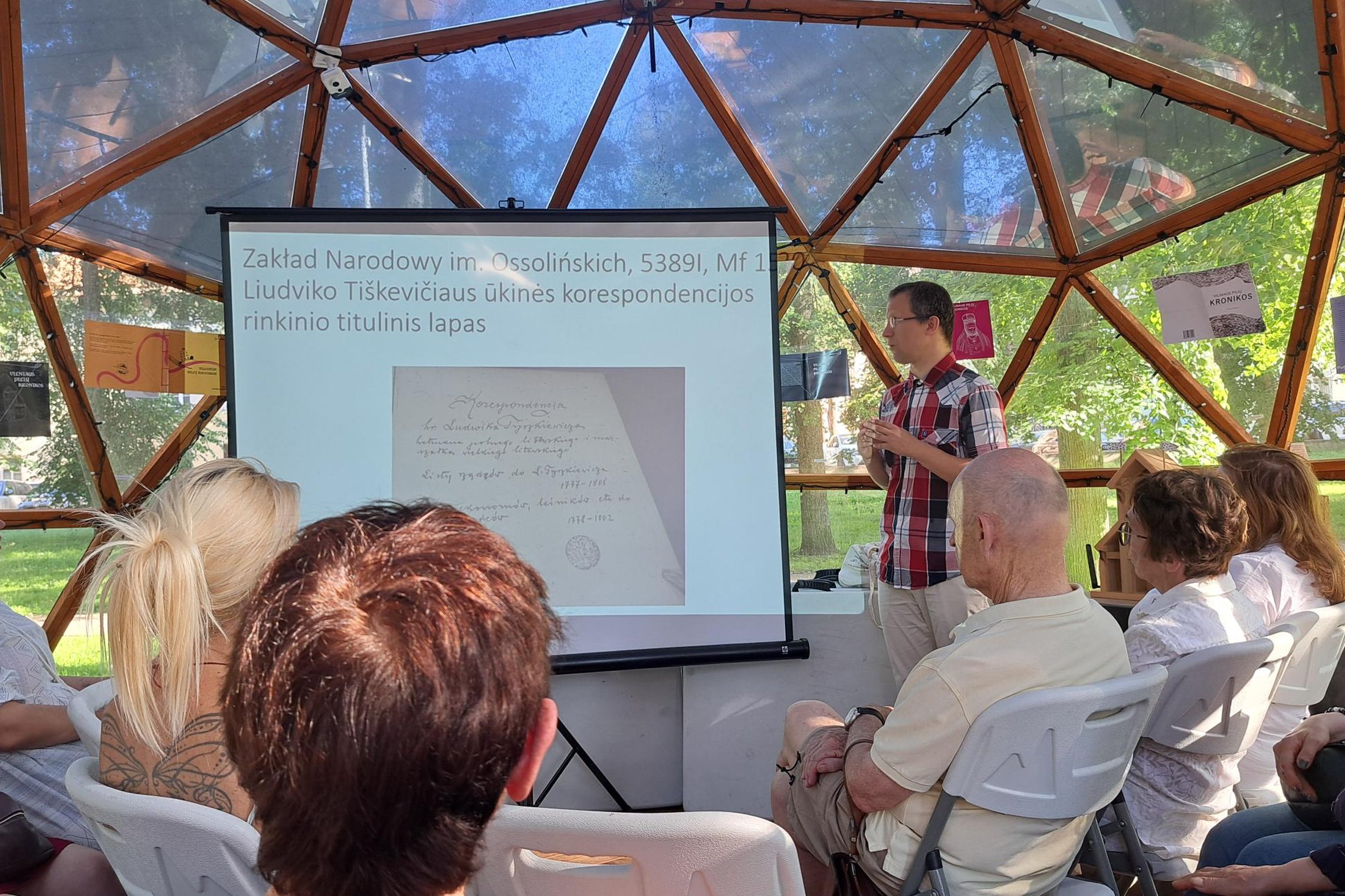Pranešėjas stovi prie projekcinio ekrano ir skaito paskaitą apie Liudviko Tiškevičiaus ūkinius dokumentus, auditorija klausosi kupolo formos patalpoje su medinėmis konstrukcijomis ir permatomomis sienomis.
