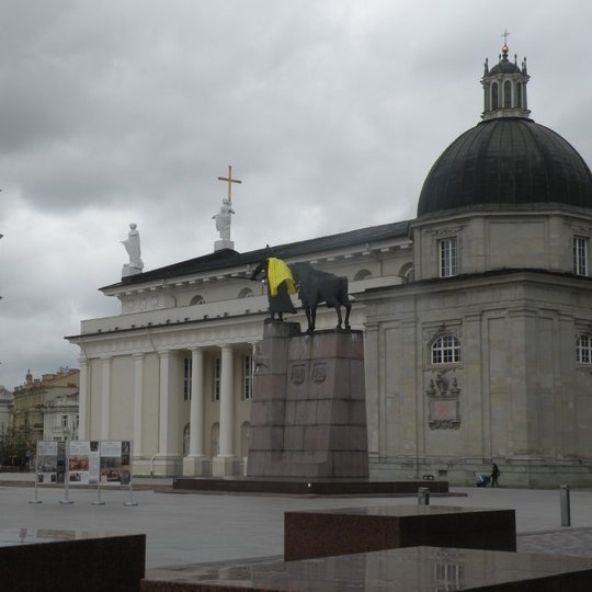 Nuotraukoje iš tolo matoma Katedros aikštė. Kairėje - aukšta varpinė, o ant jos kryžius. Žiūrint dešiniau matoma Katedros bazilika su kolonomis. Nuotraukos centre iš nugaros matoma metalinė Gedimino ir žirgo statula - paminklas ant aukšto betoninio pagrin