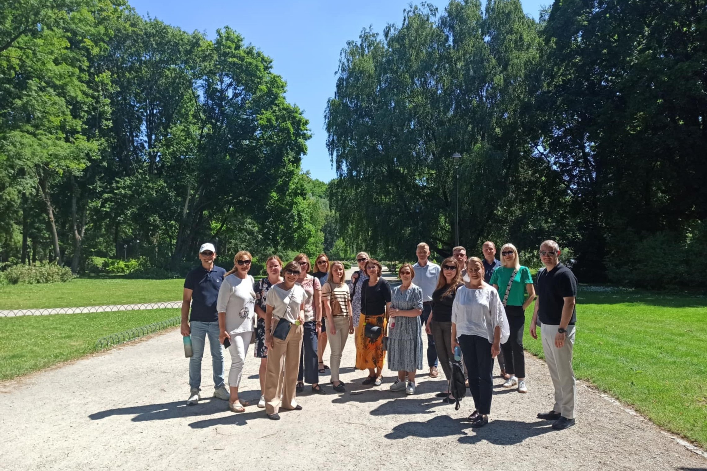 Grupė žmonių stovi saulėtame take žaliuojančiame parke. Visi dalyviai šypsosi ir pozuoja bendrai nuotraukai ekskursijos metu.