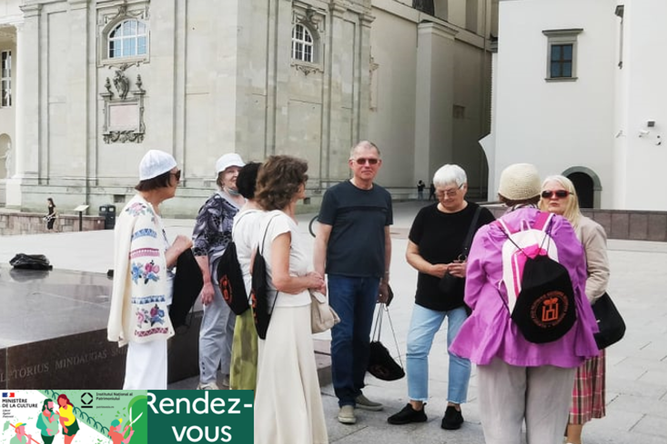Grupė ekskursijos dalyvių stovi prie Gedimino paminklo. Jie apsirengę vasariškai. Šilta diena. Už jų yra Vilniaus katedra ir Valdovų rūmai.
