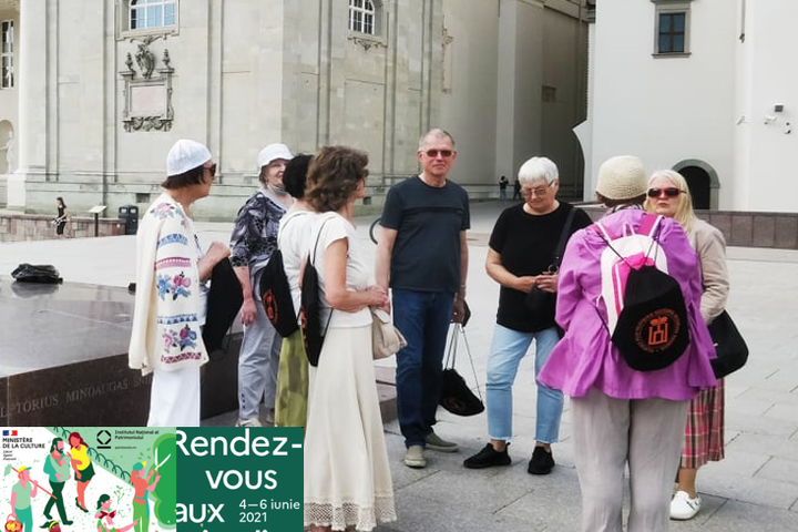 Grupė ekskursijos dalyvių stovi prie Gedimino paminklo. Jie apsirengę vasariškai. Šilta diena. Už jų yra Vilniaus katedra ir Valdovų rūmai.