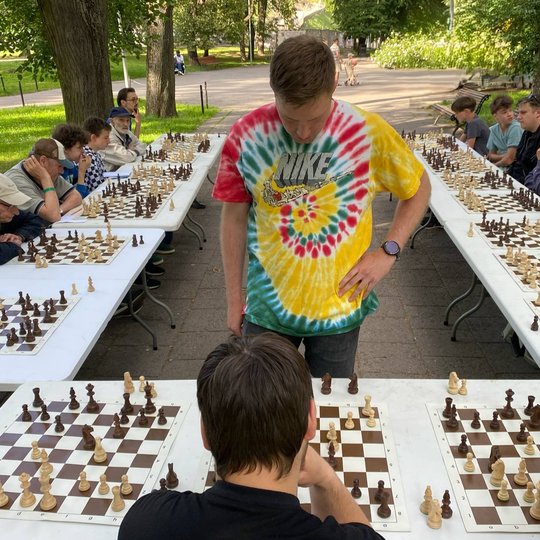 Šachmatų simultano partija po atviru dangumi: žaidėjas stovi prie šachmatų lentos, apsuptas susikaupusių dalyvių, sėdinčių prie dviejų ilgų stalų žalioje parko aplin