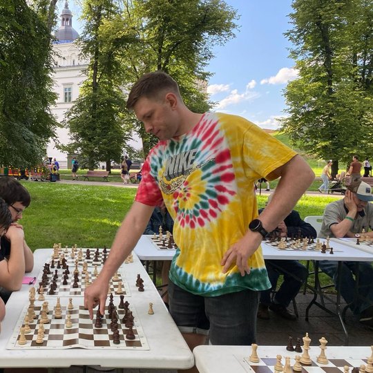 Šachmatų simultanas Vilniaus centre: žaidėjas atlieka ėjimą prieš susikaupusį jaunuolį prie lauko šachmatų lentos, fone matyti Vilniaus arkikatedros varpinė ir vaikštantys praeiviai tarp medžių.