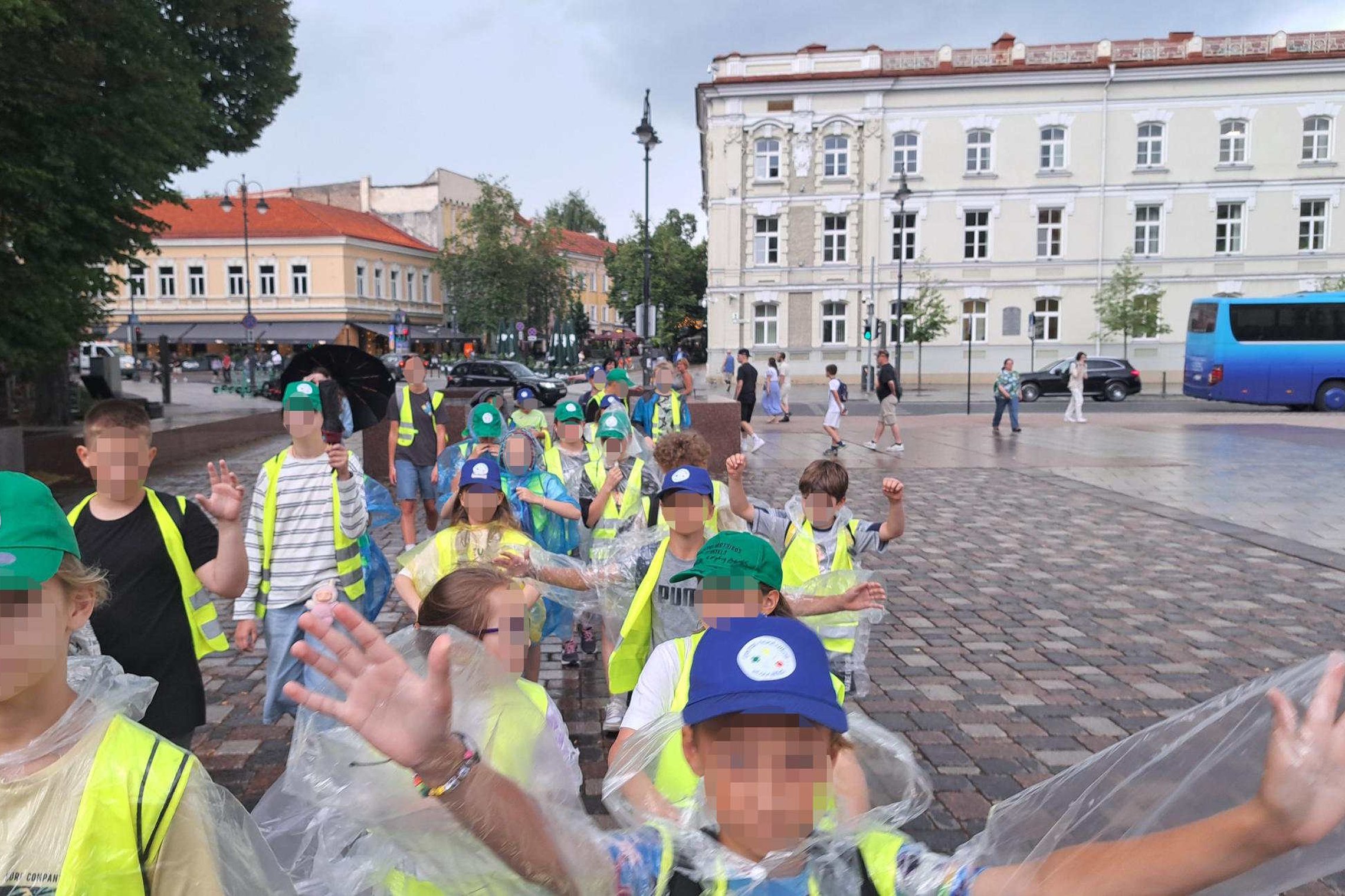 **ALT tekstas:** Vaikų grupė su ryškiaspalvėmis liemenėmis ir kepuraitėmis žygiuoja per Katedros aikštę Vilniuje, apsirengusi lietpalčiais per lietų. Fone – istorinis miesto centras su senoviniais pastatais ir šaligatviu vaikštančiais praeiviais.
