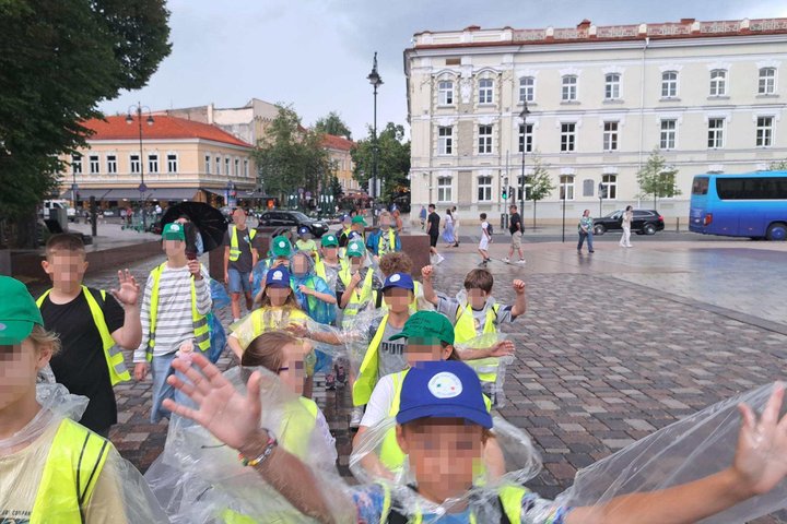 **ALT tekstas:** Vaikų grupė su ryškiaspalvėmis liemenėmis ir kepuraitėmis žygiuoja per Katedros aikštę Vilniuje, apsirengusi lietpalčiais per lietų. Fone – istorinis miesto centras su senoviniais pastatais ir šaligatviu vaikštančiais praeiviais.