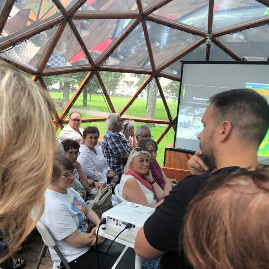 Žmonės susirinkę kupole dalyvauja paskaitoje. Priekyje stovi pranešėjas, rodomas skaidrių ekranas su žemėlapiais ir tyrimų vaizdais.