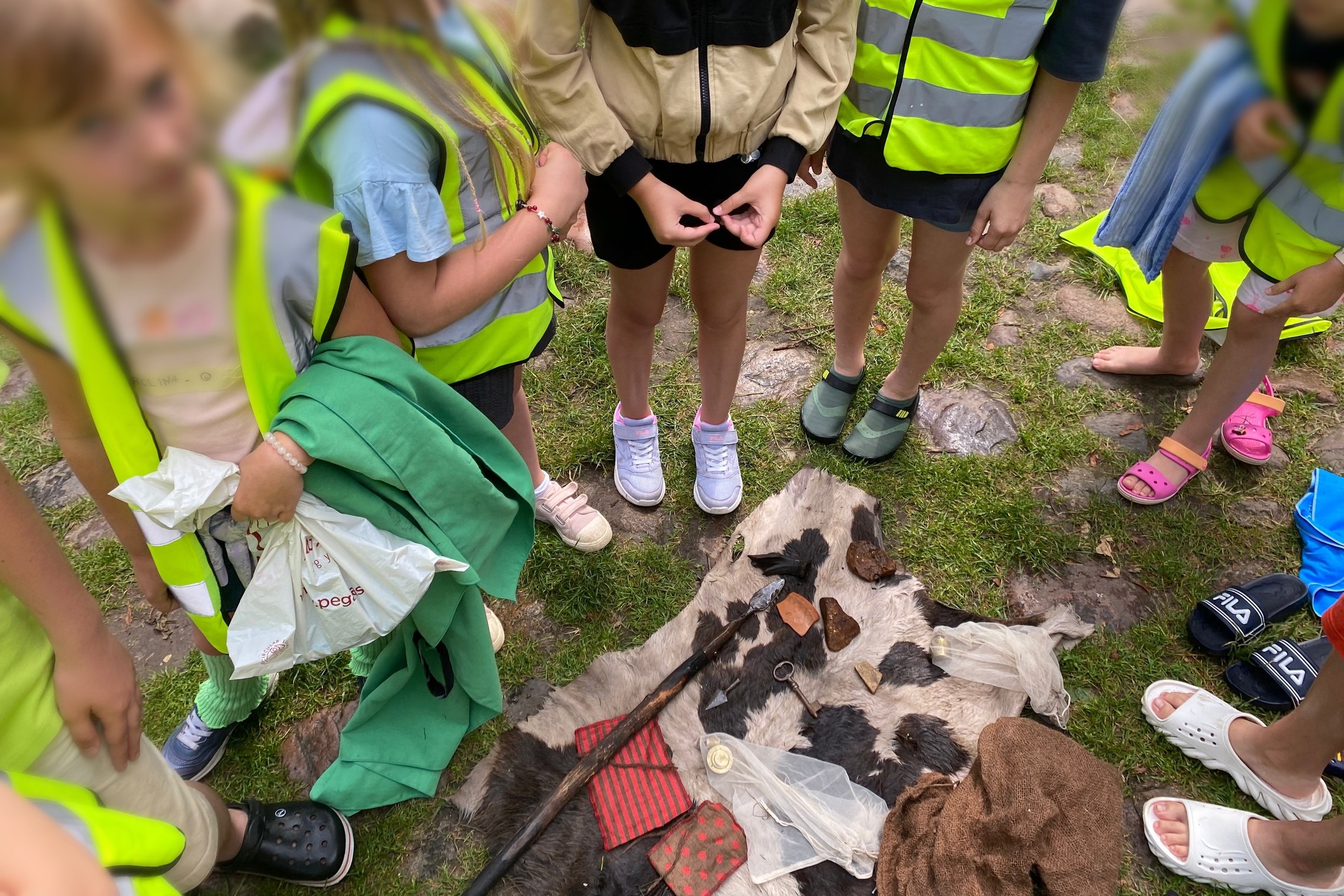 Vaikai apžiūri ant žemės išdėliotus įvairius daiktus, primenančius archeologinius atradimus, išdėliotus ant gyvūno kailio