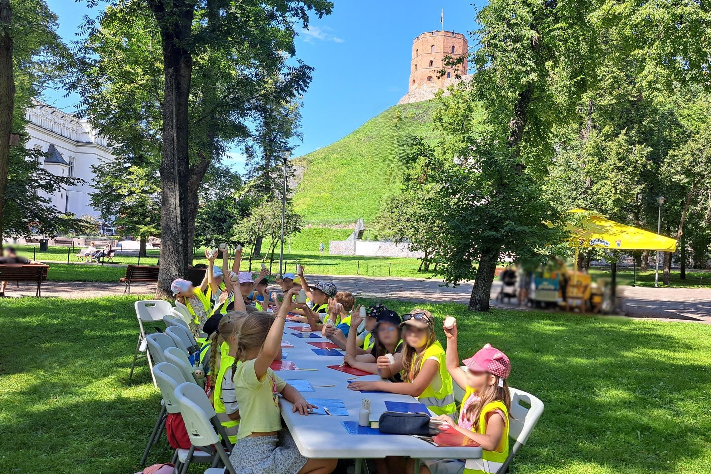 Vaikai sėdi lauke prie stalo, dalyvauja edukacijoje ir pakėlę rankas rodo savo nulipdytus molio bokštus, tolumoje matyti Gedimino pilies bokštas.