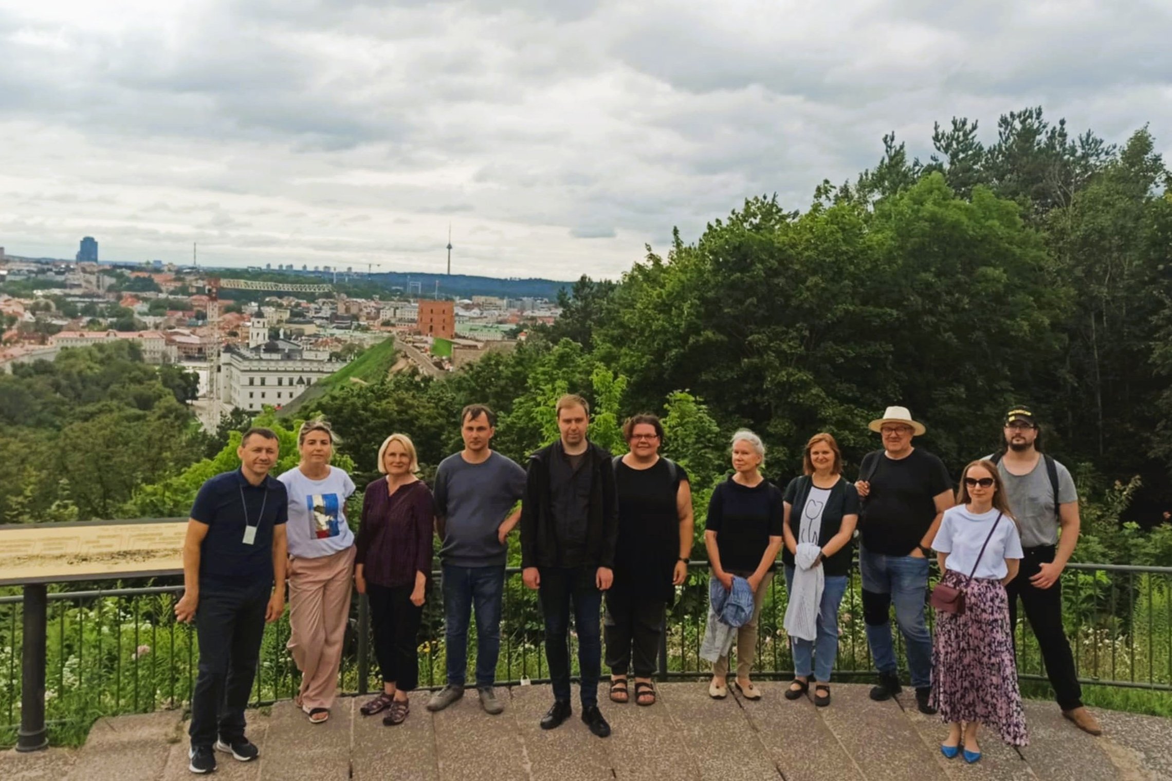 Grupė žmonių stovi ant Trijų Kryžių kalno, fone vaizdas į Vilnių. Matyti Gedimino bokštas, Valdovų rūmai, televizijos bokštas ir kiti miesto pastatai. Aikštelę supa žaluma, dangus apsiniaukęs.