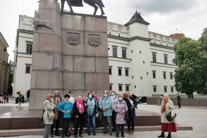 Didelė ekskursijos grupė būriuojasi prie Gedimino paminklo. Už paminklo stūkso Valdovų rūmai. Debesuota diena.