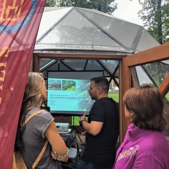 Žmonės klausosi paskaitos Vilniaus pilių kupole; fone matomas ekranas su skaidre apie Gedimino kalną, kairėje – raudona vėliava su užrašu „Vilniaus pilys“.