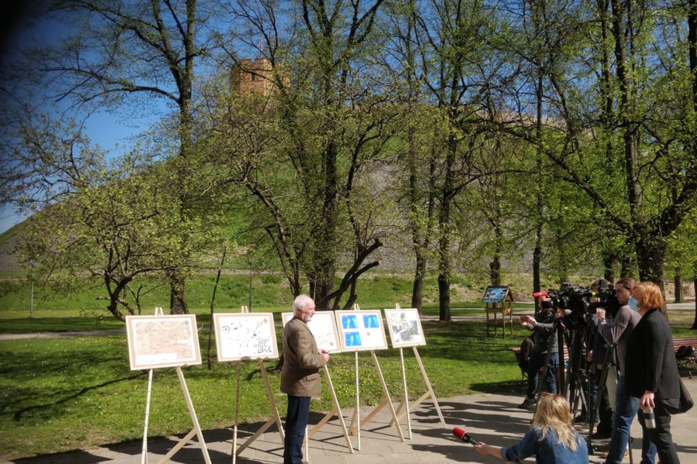 Pilių parkas, vyksta spaudos konferencija. Išrikiuoti penki molbertai su spalvotais plakatais, kuriuose vaizduojami atlikti tyrimai randant bokštą. Už molbertų medžiai, o už jų Gedimino kalnas ir Gedimino pilies bokštas. Prieš molbertus stovi žmogus. Jis 