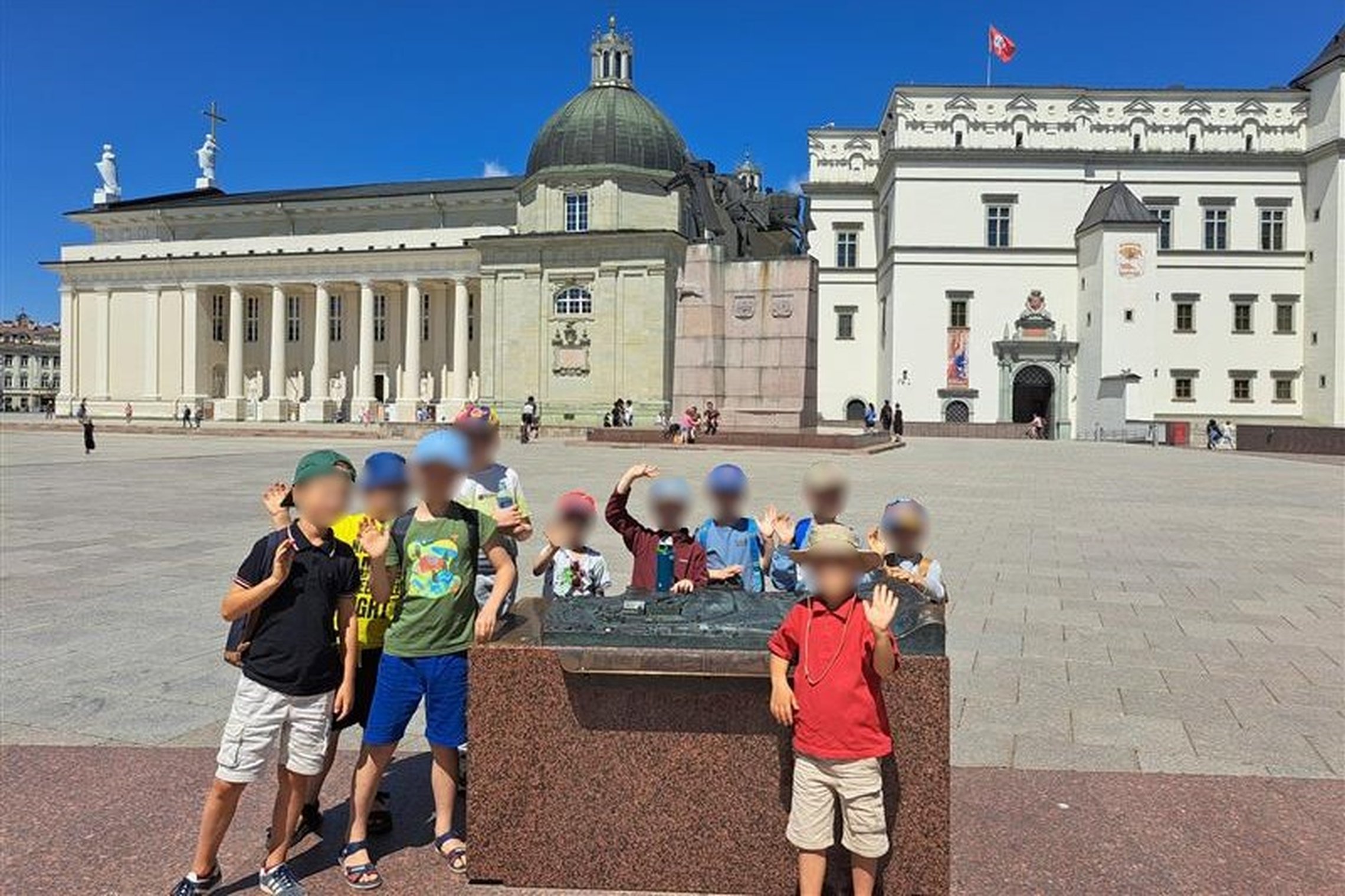 Vaikų grupė stovi prie maketo Vilniaus Katedros aikštėje, už jų matyti Vilniaus arkikatedra bazilika ir Valdovų rūmų pastatas su Lietuvos vėliava ant stogo. Vaikai mojuoja ir šypsosi saulėtą dieną.