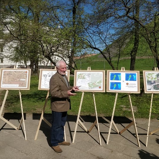 Pilių parkas, vyksta spaudos konferencija. Išrikiuoti penki molbertai su spalvotais plakatais, kuriuose vaizduojami atlikti tyrimai randant bokštą. Už molbertų medžiai, o už jų Gedimino kalnas ir Gedimino pilies bokštas. Prieš molbertus stovi žmogus. Jis 