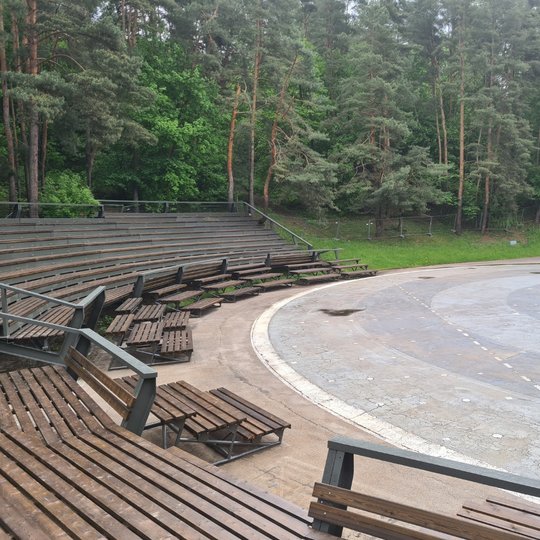 Dainų slėnio suremontuota estrada. Gražūs, nauji mediniai suolai. Yra vasara, žaliuoja medžiai.